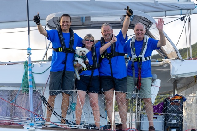 4 smiling sailors in lifejackets and crew uniforms, blue polos and blue shorts. They are waving and giving a thumbs up while the lady is holding a small white dog on a catamaran.