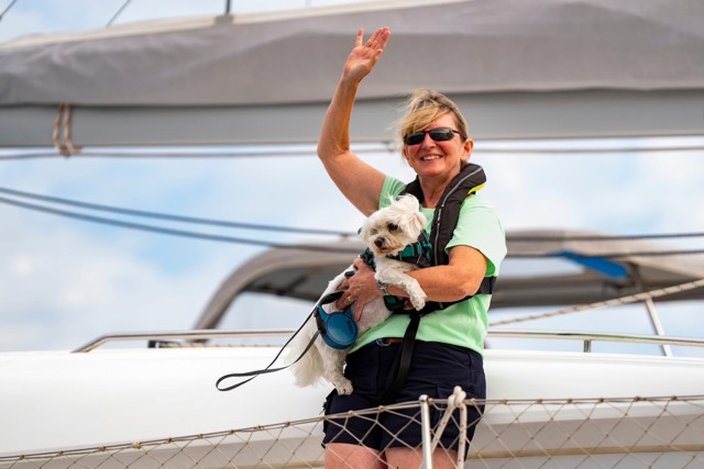 Lady in lifejacket and green t-shirt, holding small white dog on catamaran and waving.
