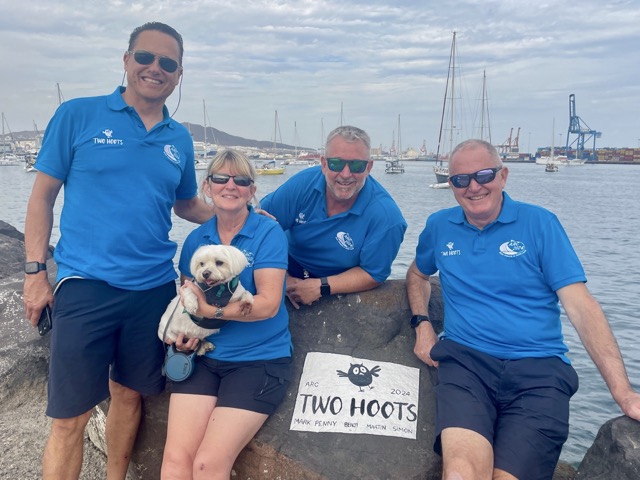 4 smiling sailors in sunglasses and crew uniforms, blue polos and blue shorts. The lady is holding a small white dog and they are all posed next to rock art reading Two Hoots.