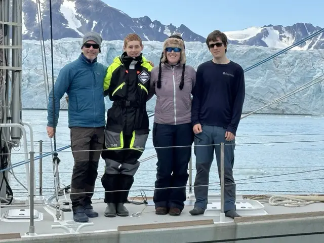4 sailors on the bow or a sailboat in front of a glacier and with mountains behind 